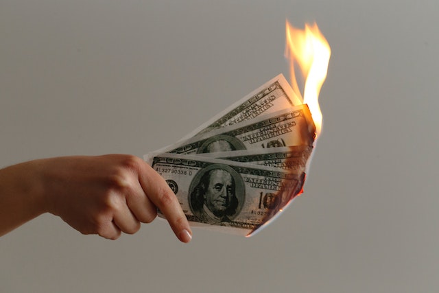 a hand holding three 100 dollar bills which are burning at the end like a cigarette
