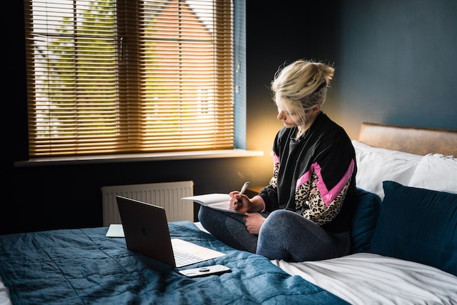 young business person in home office, writing in notebook with laptop open and phone nearby